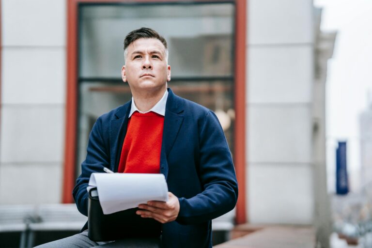 A man in a red sweater and blazer sitting outside a workplace thinks and writes by hand.