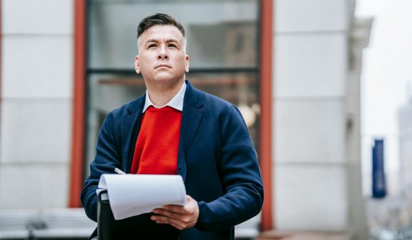 A man in a red sweater and blazer sitting outside a workplace thinks and writes by hand.